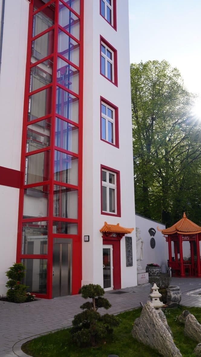 Gläserner Fahrstuhlturm mit roter Rahmenkonstruktion an der Außenwand, daneben der ebenerdige Eingang mit Pagodendach